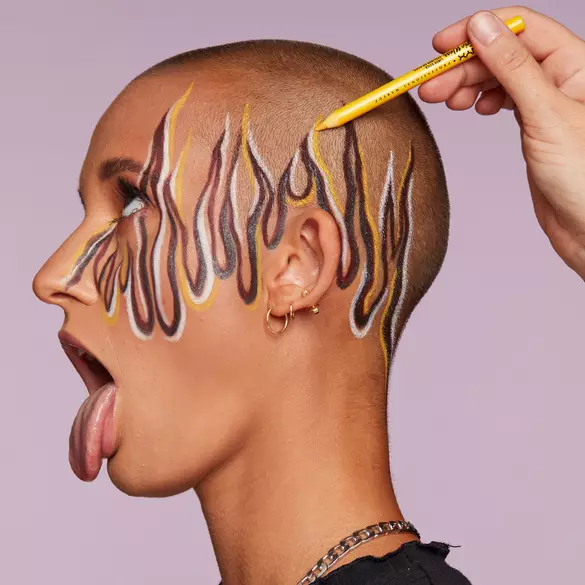 Text: Junge Frau mit rasiertem Kopf und herausgestreckter Zunge bekommt mit dem Epic Wear Liner Stick Eyeliner ein Flammenmuster auf ihren Kopf gemalt.
