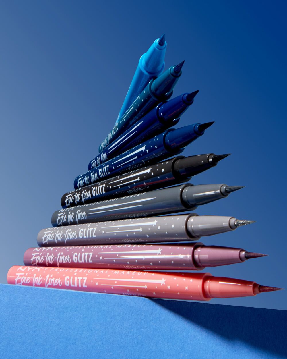 Neun ubereinander gestapelte NYC Epic Wear Lier Stix Glitz in verschiedenen Farben vor einem blauen Hintergrund