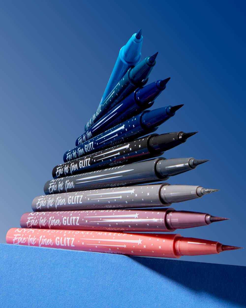 Neun ubereinander gestapelte NYC Epic Wear Lier Stix Glitz in verschiedenen Farben vor einem blauen Hintergrund