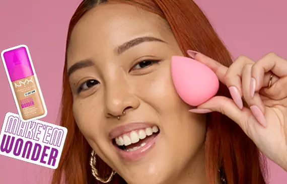 Nahaufnahme einer jungen, lachelnden Frau, die einen pinken Make-up-Schwamm in die Kamera halt neben der Make 'Em Wonder Foundation
