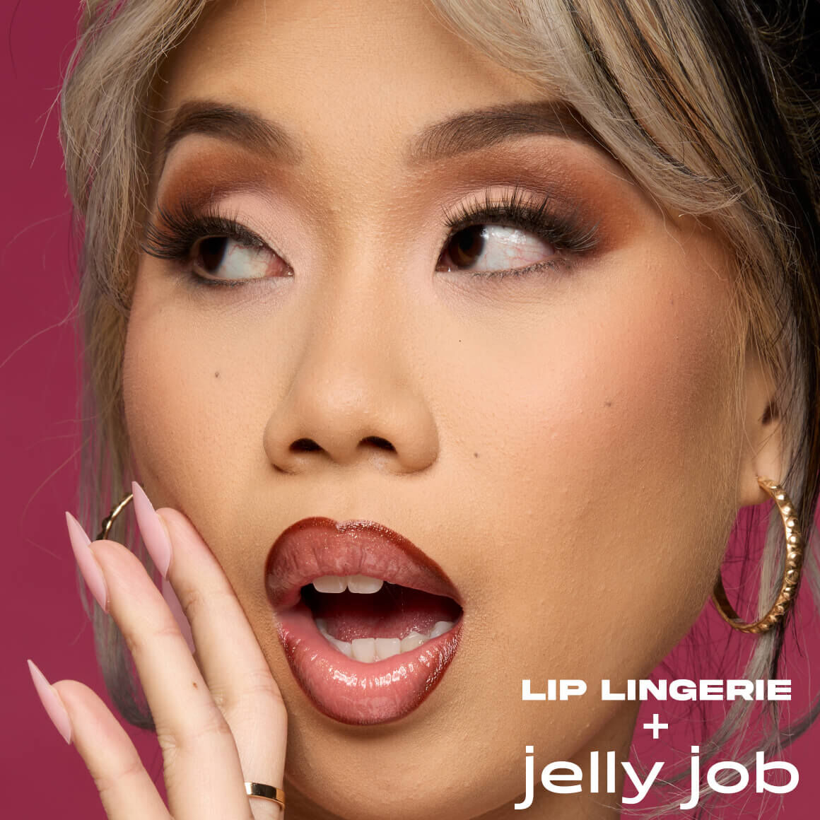 Nahaufnahme einer jungen Frau mit hellem Teint und blond gestrahntem Haar. Sie blickt mit offenem Mund und uberraschtem Ausdruck nach oben rechts und beruhrt ihr Kinn mit der Hand. Ihre Lippen sind mit einem glanzenden, dunkelroten Lippenprodukt betont, und ihre Augen sind mit braunem Lidschatten geschminkt. Sie tragt goldene Ohrringe. Auf der unteren rechten Seite des Bildes steht in weißer Schrift 'Lip LINGERIE' gefolgt von einem Pluszeichen (+) und darunter in einer geschwungenen Schrift 'jelly job'. Der Hintergrund ist magenta.