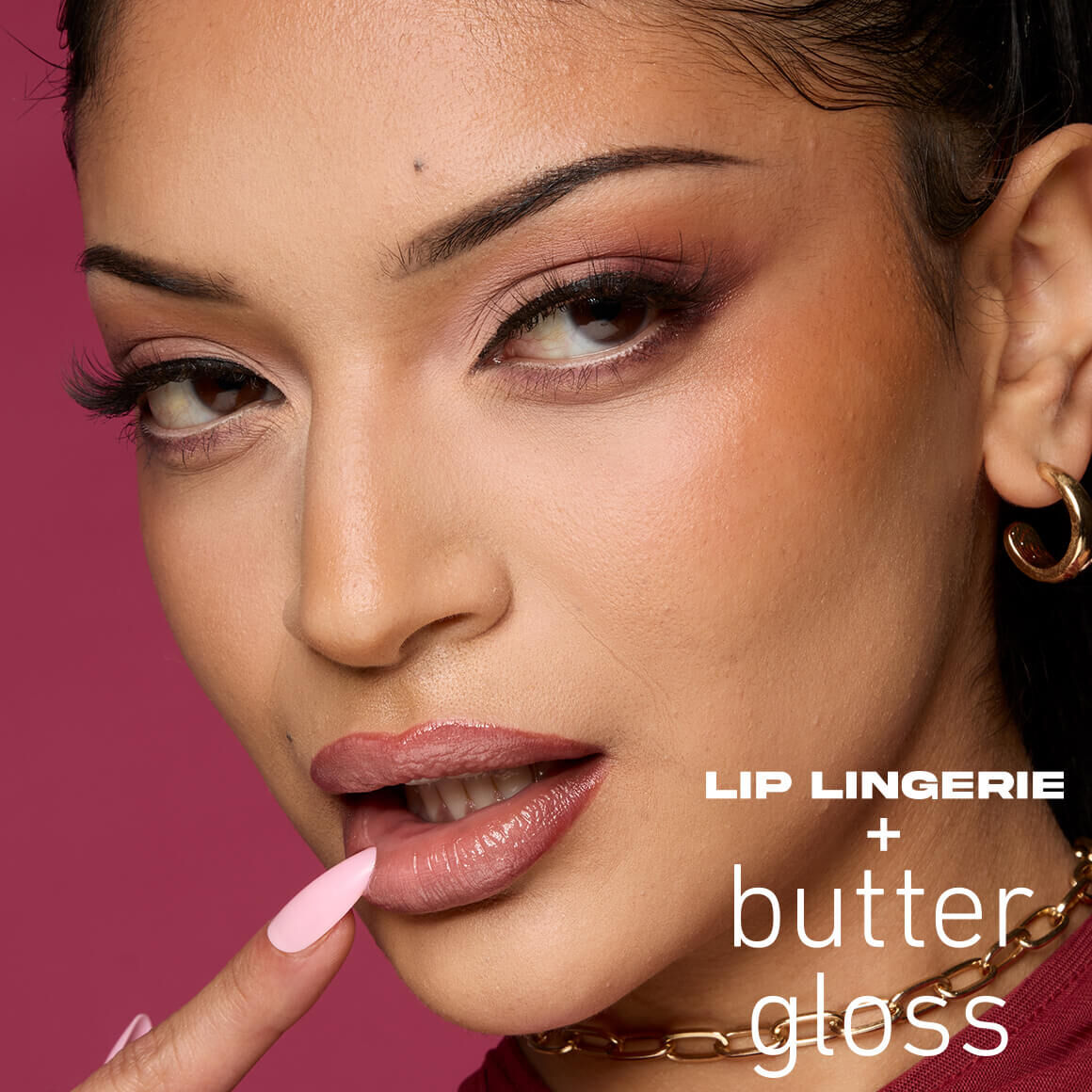 Nahaufnahme einer jungen Frau mit dunklem, welligem Haar und makellosem Make-up. Sie hat einen verfuhrerischen Blick und ihren rosafarbenen, lang lackierten Zeigefinger an ihren glanzenden, rosafarbenen Nude-Lippen. Ihre Augen sind mit einem rauchigen, rosabraunen Lidschatten und einem starken Lidstrich betont. Sie tragt goldene Creolen und eine goldene Halskette. Auf der rechten Seite des Bildes steht in weißer Schrift 'Lip LINGERIE' gefolgt von einem Pluszeichen (+) und darunter in einer geschwungenen Schrift 'butter gloss'. Der Hintergrund ist magenta.