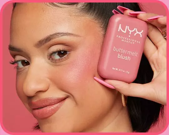 Model halt das rosa NYX Buttermelt Blush neben ihre Wange, um das frische, farbintensive Finish auf der Haut zu zeigen.