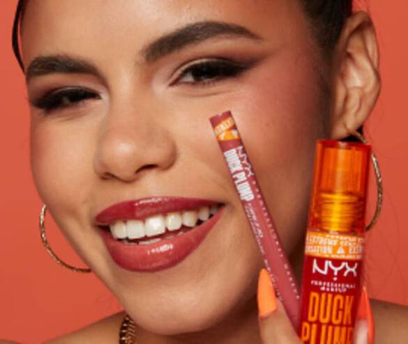 Eine Frau lachelt und halt einen Lippenstift sowie einen Lipgloss von NYX in der Hand, vor einem orangefarbenen Hintergrund.