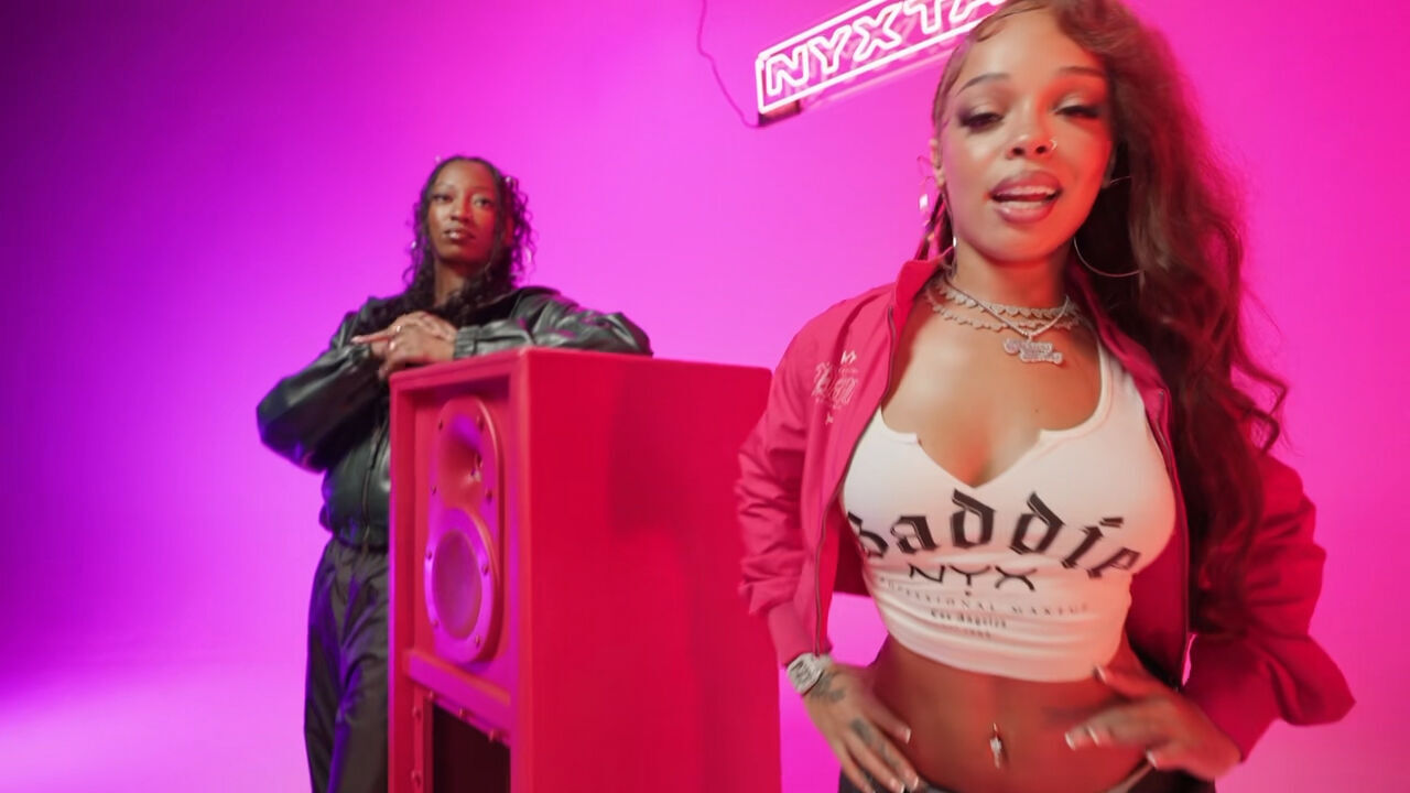 NYX Professional Makeup „NYXTAPE“-Musikvideoszene mit Wondagurl und Honey Bxby neben einem roten Lautsprecher in einem pink beleuchteten Studio, daruber ein leuchtendes „NYXTAPE“-Neon-Sign.