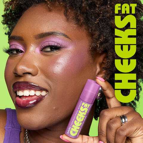 Frau mit dunklem Hautton tragt Fat Cheeks Juicy Blush von NYX Professional Makeup auf Wangen und Augenlidern und halt den Liquid Blush zwischen Daumen und Zeigefinger neben ihrem Gesicht