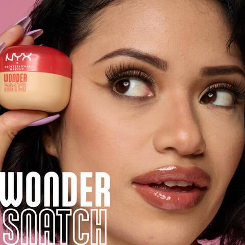 Nahaufnahme einer Frau mit Make-up, die ein kleines, beiges Produkt mit rotem Deckel in der Hand halt, auf dem "NYX PROFESSIONAL MAKEUP WONDER SNATCH" steht. Die Frau hat lange, volle Wimpern, definierte Augenbrauen und glanzende Lippen. Auf dem Bild sind außerdem in großen, weißen Umrissbuchstaben die Worte "WONDER SNATCH" zu sehen.