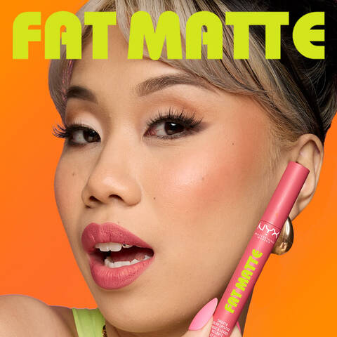 Frau halt den NYX Fat Matte Lippenstift in einem matten Mauve-Ton an ihren Kiefer, vor leuchtend orangen Hintergrund.